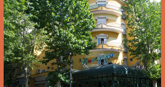 Edificio giallo con balconi e alberi verdi davanti.