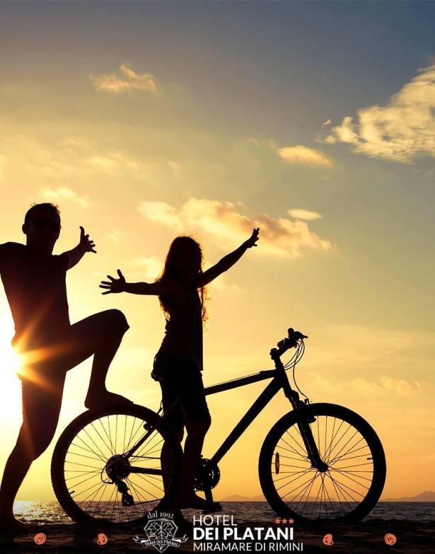 Zwei silhouettierte Personen mit Fahrrad bei Sonnenuntergang am Strand.